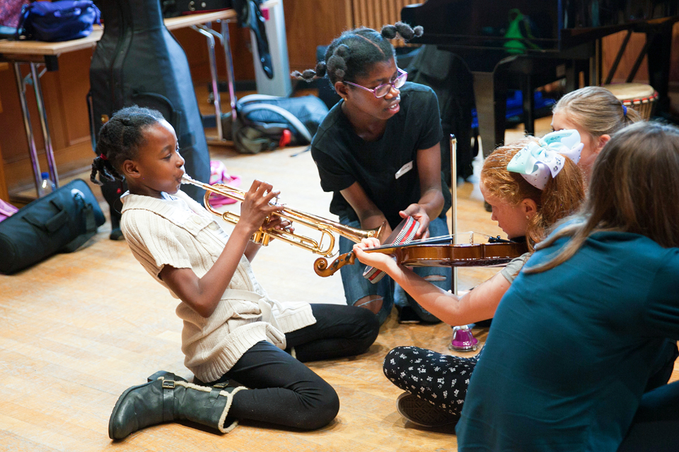 A black girl playing the trumpet, with a white girl with ginger hair, playing the violin opposite her, with other students around them.